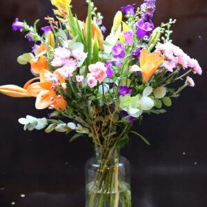 Zomerpalet Boeket met oranje lelies, paarse klokjesbloemen en gele celosia in een cilindrische glazen vaas op een houten krat
