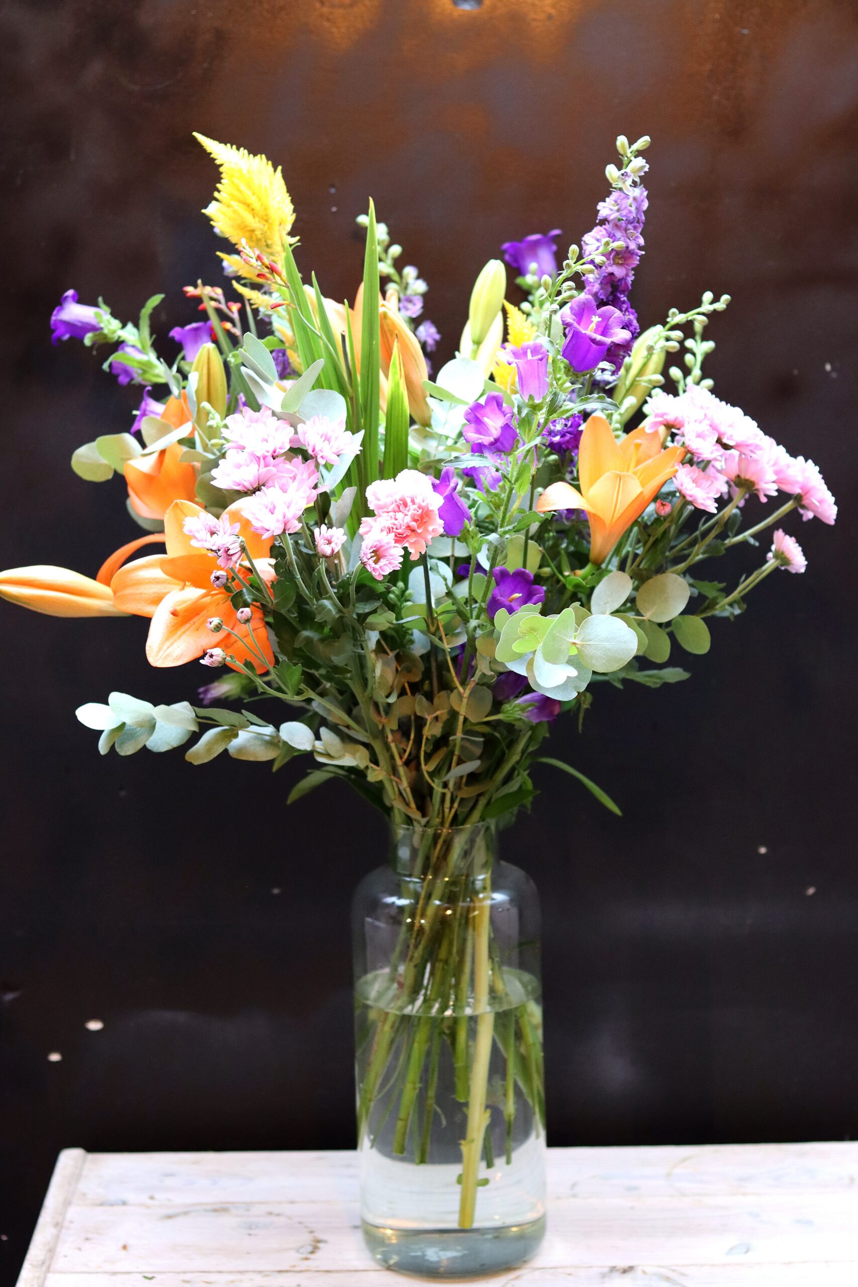 Zomerpalet Boeket met oranje lelies, paarse klokjesbloemen en gele celosia in een cilindrische glazen vaas op een houten krat
