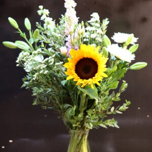 Zonnestraal Boeket met een grote zonnebloem, witte chrysanten en roze violieren in een glazen vaas op een houten krat