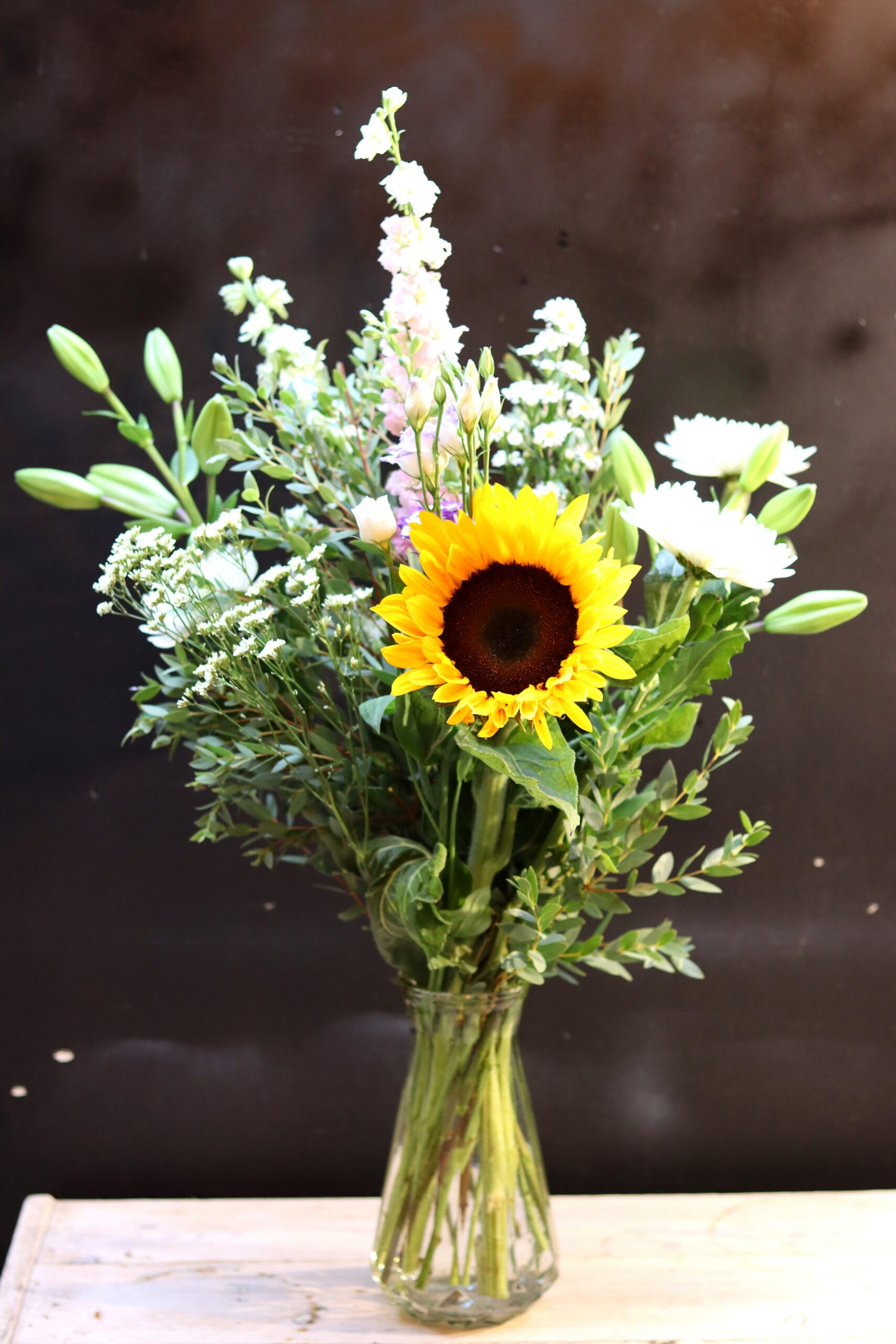 Zonnestraal Boeket met een grote zonnebloem, witte chrysanten en roze violieren in een glazen vaas op een houten krat